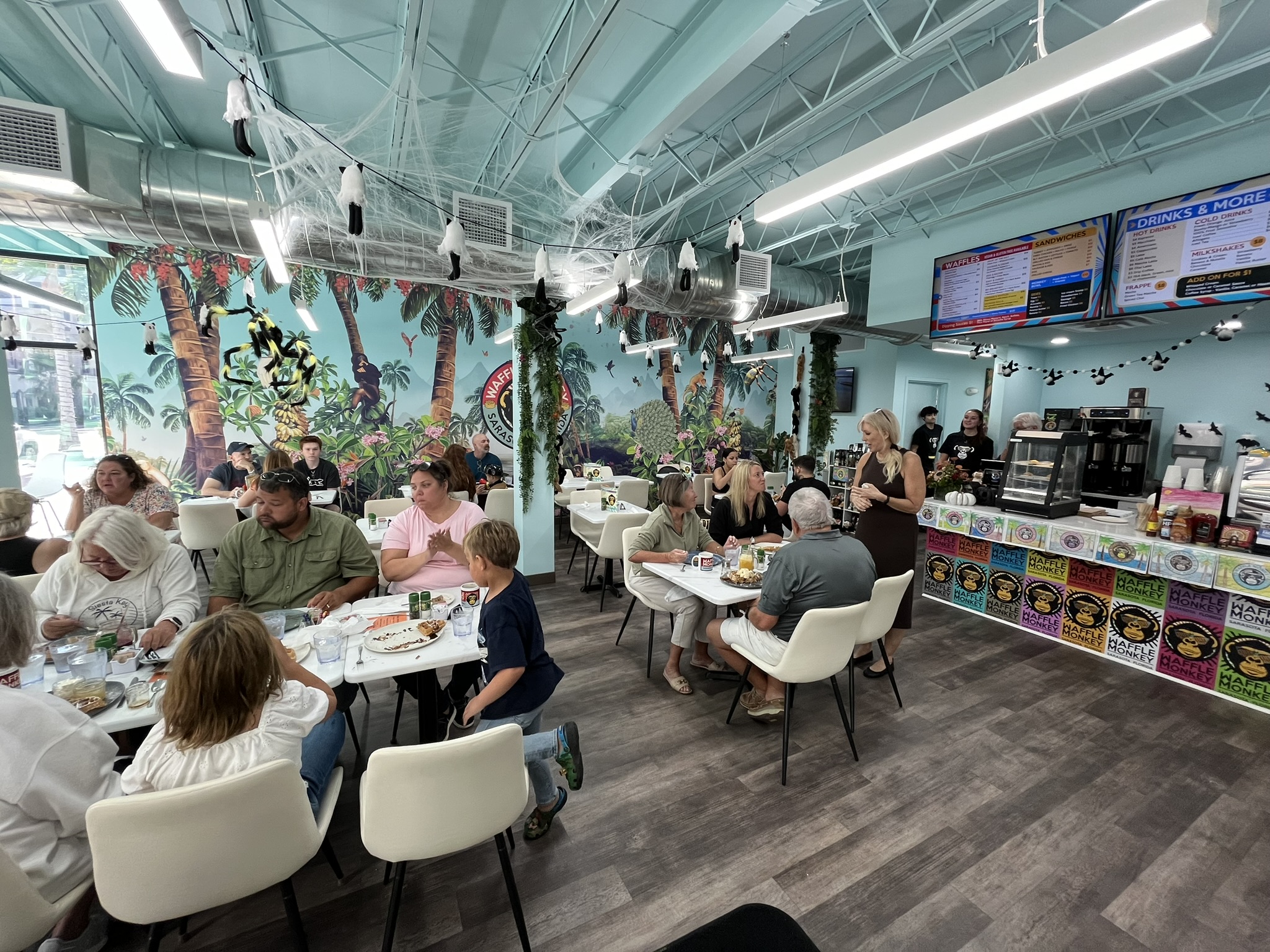 A busy evening inside the Waffle Monkey Sarasota cafe — tables of guests, the tropical mural and the coffee counter